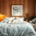 A cozy, well-lit bedroom with a plush, oversized bed dressed in soft, neutral linens. Warm, ambient lighting from a brass floor lamp casts a gentle glow, while a large, framed abstract art piece hangs above the headboard. A cozy, tufted armchair sits in the corner, inviting relaxation, with a fluffy, grey throw draped over the back. The room is accented with natural wood tones, potted plants, and textural details like a woven basket and sheer curtains that drift in the breeze, creating a serene, calming atmosphere.